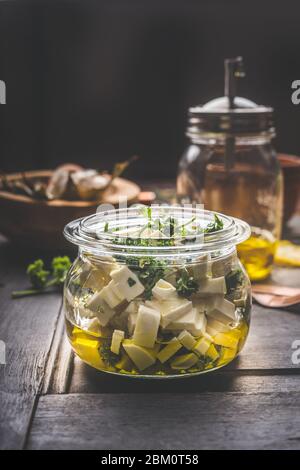 Homemade feta cheese cubes in jar with olives oil and herbs on dark rustic table background.  Marinated feta cheese. Tasty food. Country style. Stock Photo