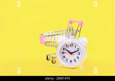 Mini shopping cart and white alarm clock on yellow background. Conceptual image of sale, seasonal discounts in shopping stores, discount time, Time sa Stock Photo