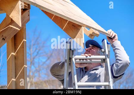 Senior gray-haired builder collects the frame of a wooden country house standing on the stairs against the blue sky. The physical activity of the elde Stock Photo