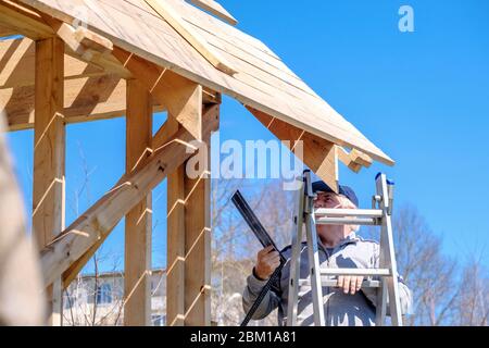 Senior gray-haired builder collects the frame of a wooden country house standing on the stairs against the blue sky. The physical activity of the elde Stock Photo