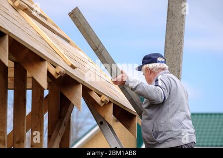 Senior gray-haired builder collects the frame of a wooden country house standing on the stairs against the blue sky. The physical activity of the elde Stock Photo