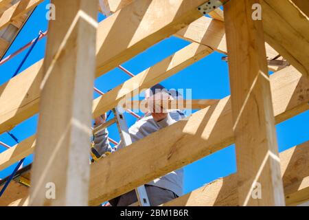 Senior gray-haired builder collects the frame of a wooden country house standing on the stairs against the blue sky. The physical activity of the elde Stock Photo