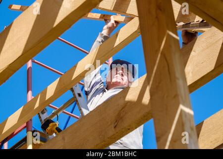 Senior gray-haired builder collects the frame of a wooden country house standing on the stairs against the blue sky. The physical activity of the elde Stock Photo