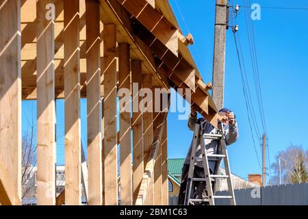 Senior gray-haired builder collects the frame of a wooden country house standing on the stairs against the blue sky. The physical activity of the elde Stock Photo