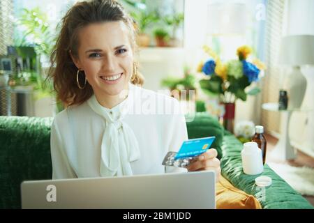 smiling young woman in white blouse with credit card and empty blister pack of pills ordering medicine online on a laptop in the modern house in sunny Stock Photo