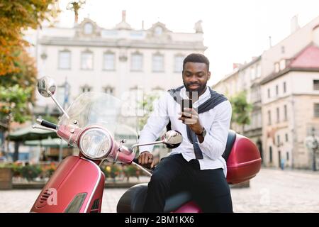 Portrait of smiling young bearded african american guy in smart casual wear using his smartphone, while sitting on the red scooter outdoors on the old Stock Photo