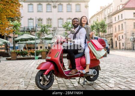 Young beautiful multiracial couple, African guy and Caucasian girl, riding on red motorbike on the city street, happy woman holds shopping bags Stock Photo