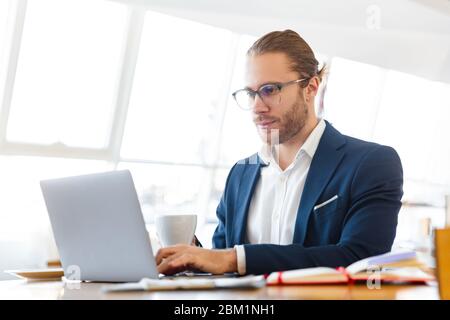 Image of handsome concentrated young businessman indoors in office using laptop computer. Stock Photo