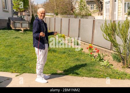 Warming up before workout. Old man in stylish sportswear is jumping with a skipping rope on the outdoor sports ground at home. Stock Photo