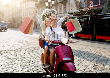 Happy smiling pretty girl with shop bags in her hands sitting behind handsome man on the red scooter and riding through the city street. Urban Stock Photo