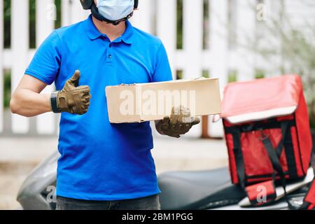 Delivery man with cardboard box standing at his scooter and showing thumbs-up Stock Photo