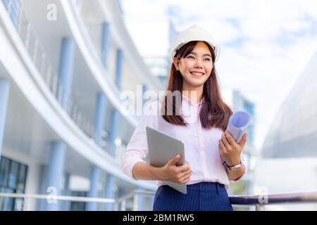Happy young attractive female Asian engineer wearing hard hat holding her computer tablet and blueprint while standing outside of office building, bus Stock Photo