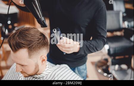 Close-up master Barber does hairstyle and styling with dryer hair to guy. Concept Barbershop Stock Photo