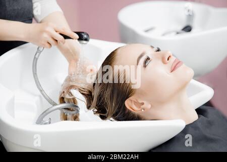 Concept beauty salon. Hairdresser washes hair of beautiful blonde girl under tap in wash Stock Photo