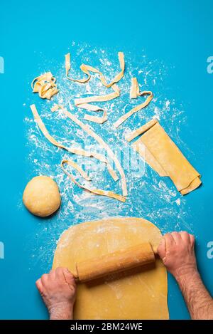 Man rolling semolina dough to make pasta, on a blue background. Top view of making tagliatelle pasta. Authentic Italian food recipe. Preparing dinner. Stock Photo