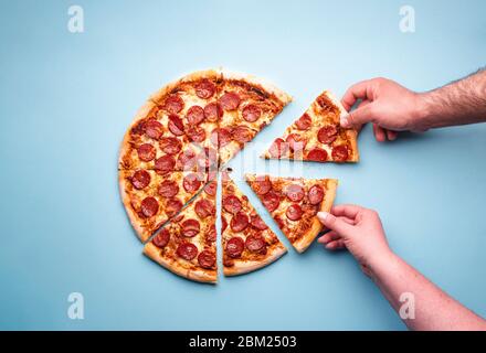 Man and woman hands grabbing pizza slices. Sliced pepperoni pizza on a blue background, above view. Top view with delicious pepperoni pizza. Stock Photo