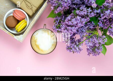 Cup of black coffee with milk and gift, lilac flowers and sweet pastel french macaroons on pink table. Top views, close-up Stock Photo