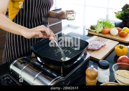 The housewife dressed in an apron putting butter in the pan prepared for frying steaks. Stock Photo