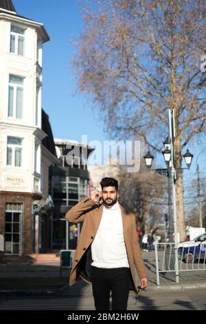 Portrait of fashionable well dressed bearded man at street. Young Indian man in brown coat talking by phone. Stock Photo