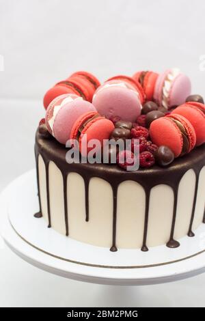 Cake with melted chocolate, red and pink french macaroons, fresh raspberry and chocolate truffles on white cakestand. Plain background Stock Photo