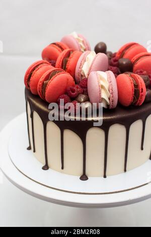 Cake with melted chocolate, red and pink french macaroons, fresh raspberry and chocolate truffles on white cakestand. Plain background Stock Photo