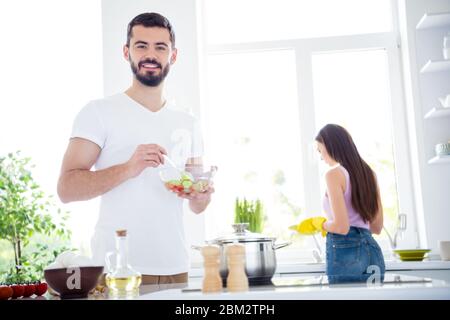 Idyllic family portrait concept. two people stay home kitchen man mix salad spoon bowl his wife wash plate in yellow rubber latex gloves sponge after Stock Photo