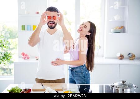 Portrait of two people spouses chef prepare organic meal dish man hold cherry tomato close cover eyes face woman humor laugh enjoy in house kitchen Stock Photo