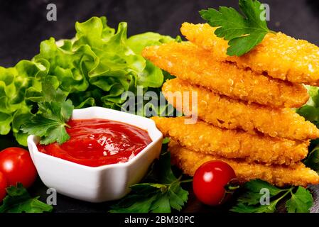 Fast homemade food.Chicken breaded nuggets on a black kitchen plate with ketchup and basil leaves, lettuce and tomatoes on a wooden background. Stock Photo