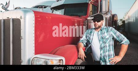 Semi truck professional driver on the job in casual clothing. Confident looking trucker stands next to red big rig wearing sunglasses and baseball cap Stock Photo