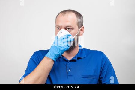 Portrait of man in his mid 30's equipped with protective mask and surgical mask on white. Healthcare worker holds hand over face mask. Stock Photo