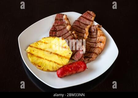The pieces of meat are juicy, chopped, with pineapple and sausage on a white plate. Dark black background. Close-up. Stock Photo