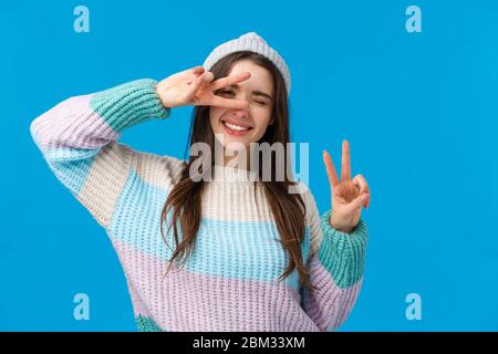 Pack your things and travel. Joyful and carefree upbeat smiling woman, wear winter sweater and hat, wink camera and make peace signs, tilt head Stock Photo