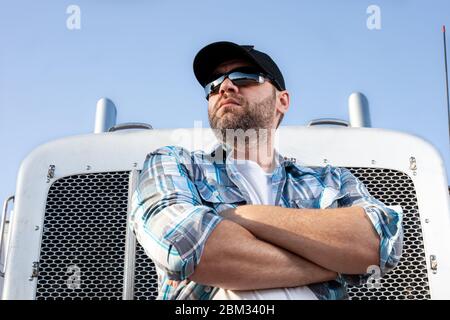 Confident semi truck driver wearing plaid shirt and black baseball cap  stands with arms crossed in front of big rig. Stock Photo