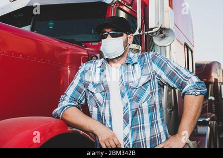semi truck professional driver on the job in casual clothing wears safety medical face mask. Confident looking trucker stands next to red big rig Stock Photo