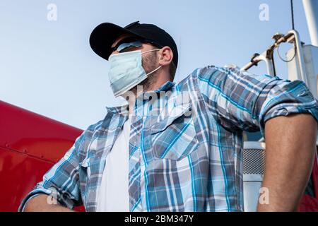 Cool looking truck driver dressed in casual clothing with plaid shirt and black baseball cap wears protective medical face mask. Stock Photo