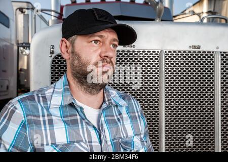 Worried semi truck driver wearing plaid shirt and black baseball hat sits in front of big rig. American trucker Stock Photo