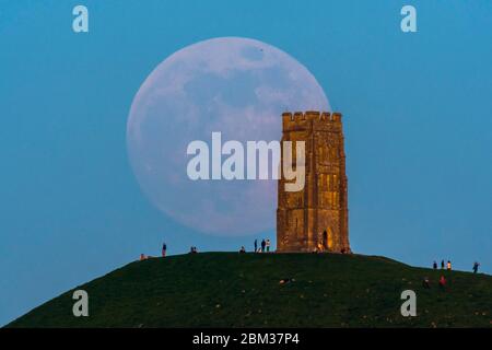 Glastonbury, Somerset, UK.  6th May 2020.  UK Weather.  The almost full moon rises up behind Glastonbury Tor in Somerset shortly before sunset with people on the top of the hill watching.  This months moonrise is called the flower moon and is going to be a supermoon.  Picture Credit: Graham Hunt/Alamy Live News Stock Photo