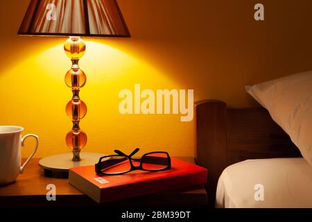 Pair of glasses upon a book on a bedside table by a bed. Stock Photo