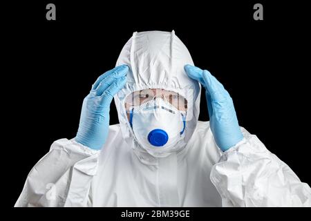 Man in white protective suit holds his head. Man in facepiece respirator and glasses, hands in disposable blue gloves, isolated on black background Stock Photo