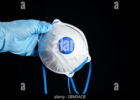 Filtering facepiece respirator in hand in blue glove, isolated on black background. N95 respirator. White FFP2 respirator, protection against coronovi Stock Photo