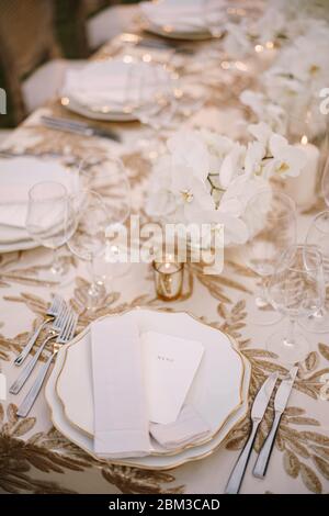 Wedding dinner table reception. Delicate creamy pastel tones of tablecloths and plates on table, three forks left and two knives on right, glasses and Stock Photo