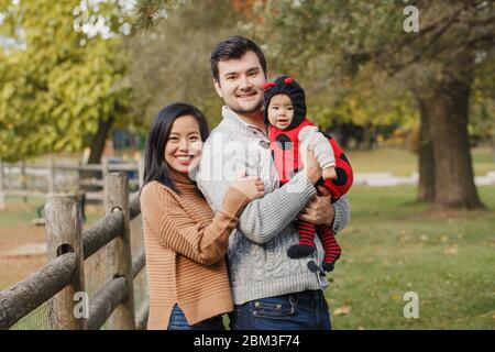 Happy smiling Asian Chinese mother and Caucasian father dad with baby girl in ladybug costume. Family in autumn fall park outdoor. Halloween or Thanks Stock Photo