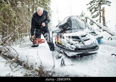 Retired man using chainsaw to clear trails while snowmobiling. Stock Photo
