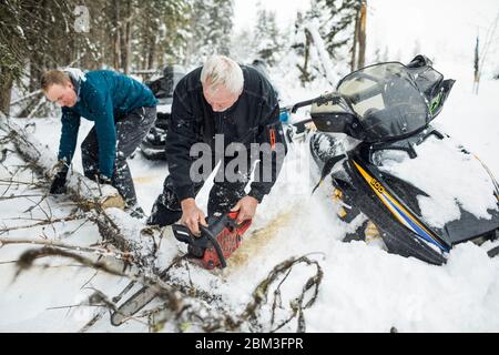 Elderly man and son using chainsaw to clear trails while snowmobiling. Stock Photo