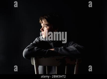Low key portrait of adolescent boy sitting on a chair in a dark room. Stock Photo
