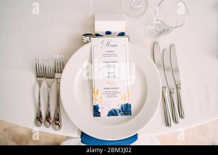Wedding dinner table reception. White plate on the table, three forks on the left, three knives on the right. The wedding menu is in the plate. Top Stock Photo