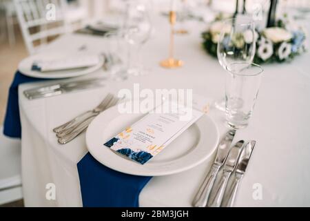 Wedding dinner table reception. White plate on the table, three forks on the left, three knives on the right. The wedding menu is in the plate. Top Stock Photo