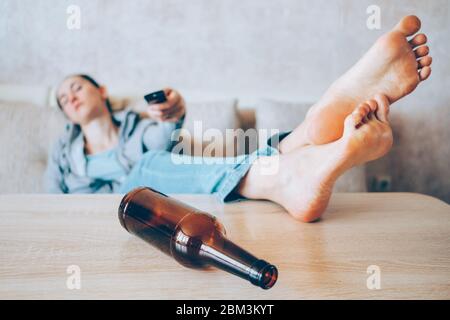 A drunk girl sits on a couch and switches the channel of the TV pultomot with a bottle in focus. Close up. Stock Photo