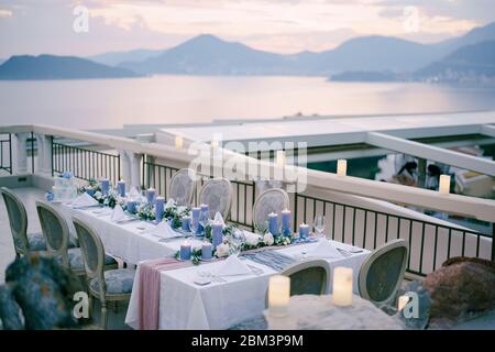 Wedding dinner table reception with lots thick blue candles against sunset sky over the mountains and sea in Budva, Montenegro. Old soft chairs on the Stock Photo