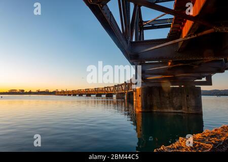 Sunrise across Tauranga harbour catches historic railway bridge as it disappears across the bay, New Zealand.